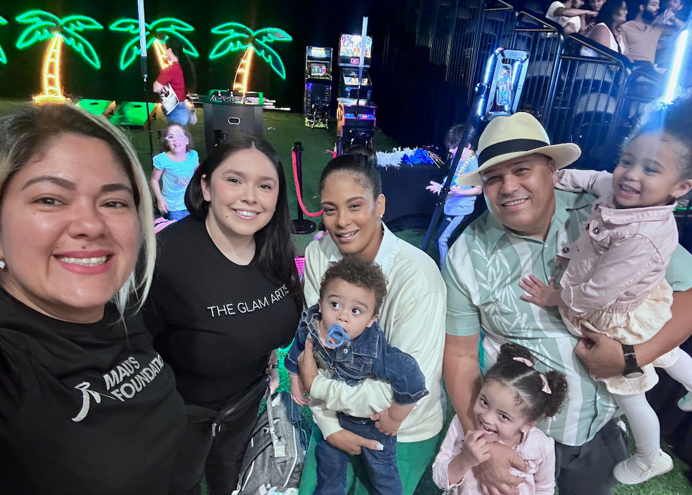 A group of six people posing for a photo at an outdoor event. Includes adults holding babies and lit-up palm tree decorations in the background.