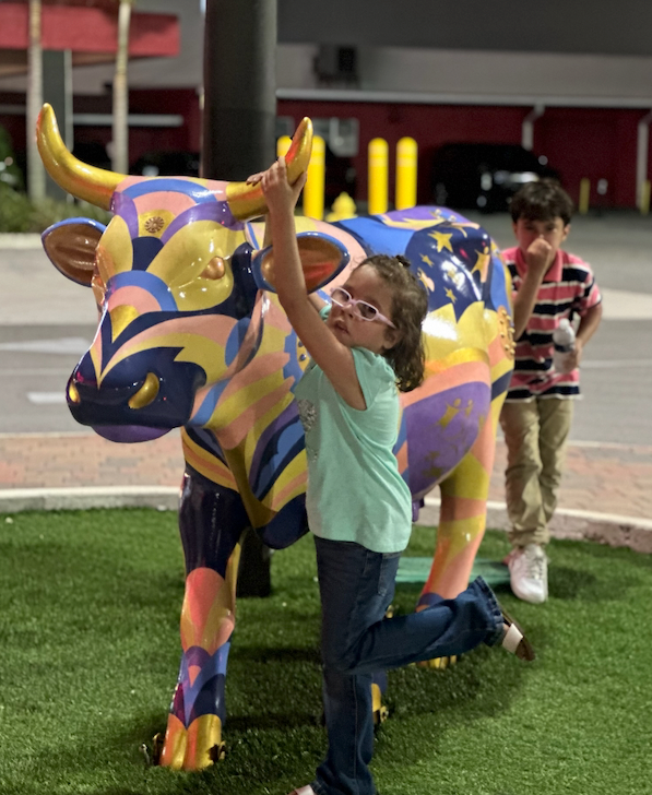 Girl in sunglasses poses with a colorful cow statue, raising her arm. A boy looks on in the background. Outdoors, near a building.