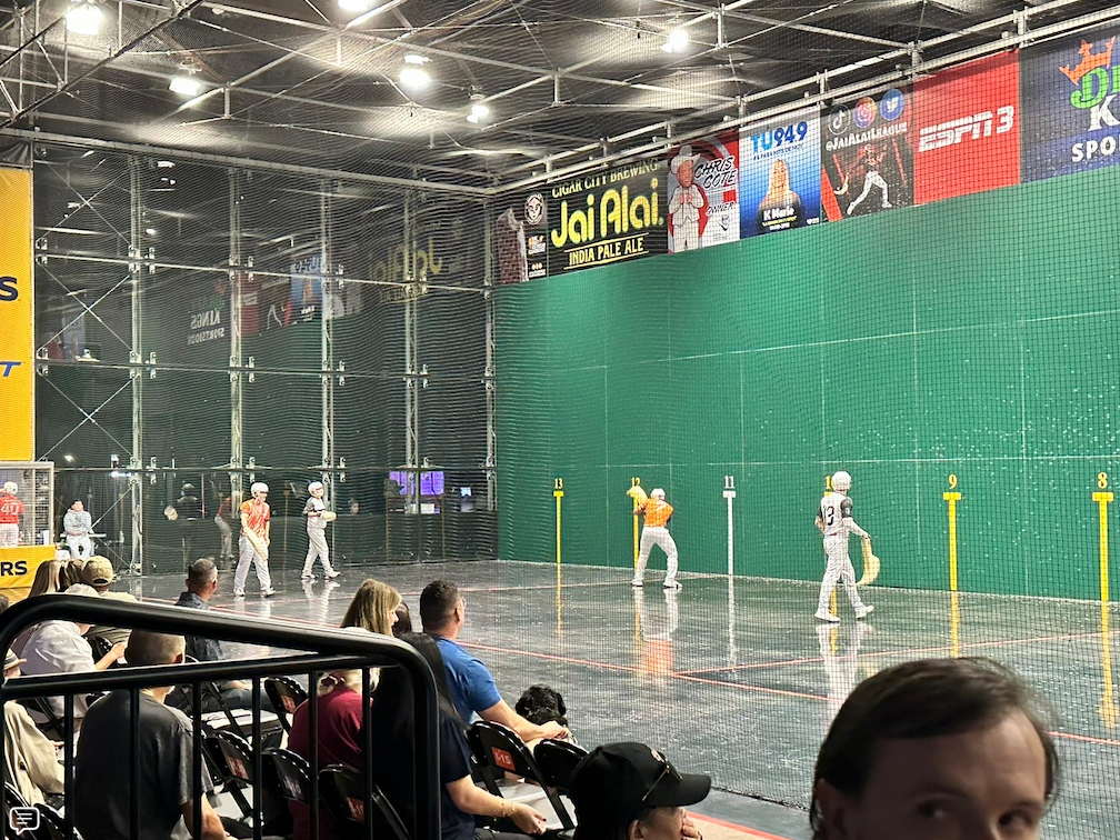 Jai alai players on a court with a green wall. Spectators watch from behind a fence.