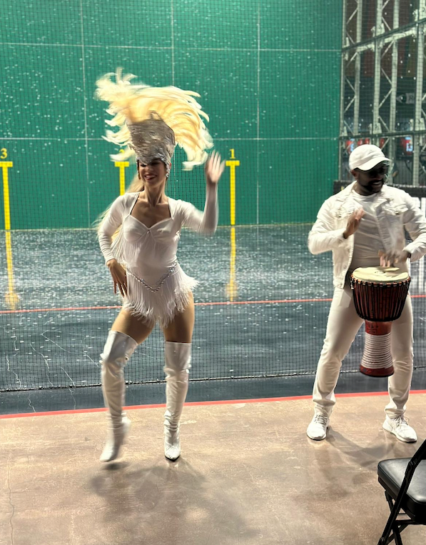 Woman in white feathered costume dances, man plays drum; both wear white, in a green-walled space.