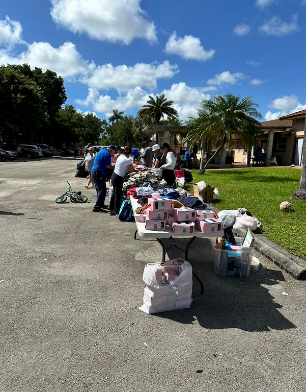 People organizing items on tables outdoors; donation event in a parking lot with clothes, toys, and gift boxes.