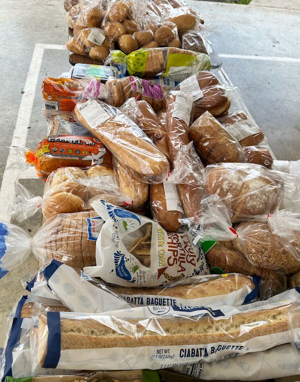 A table overflowing with various loaves of bread, including baguettes, rolls, and sliced loaves, likely for donation or distribution.