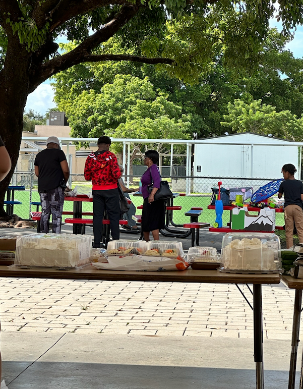 People gather at an outdoor event with tables of food. A man in a red jacket stands near a woman in a purple top.