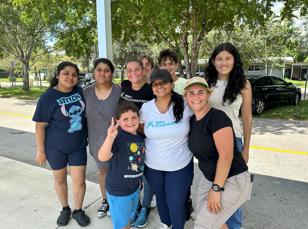 Group of people of various ages smiling and posing outdoors, near a building and trees.