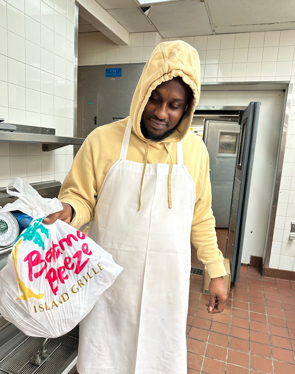 A man in a yellow hoodie and white apron holds a bag from Bahama Breeze in a kitchen. He smiles slightly.