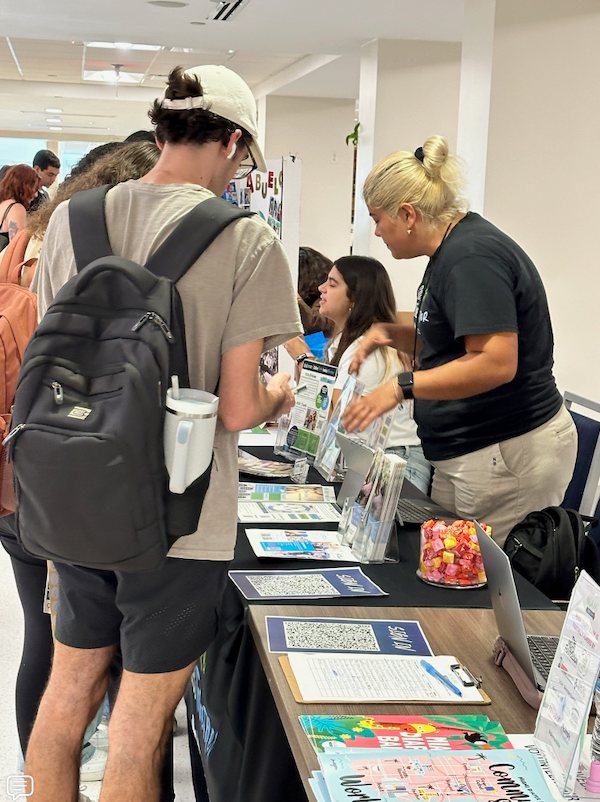 People gather around a table at an event. A woman speaks with a student, while others look at brochures and other materials.