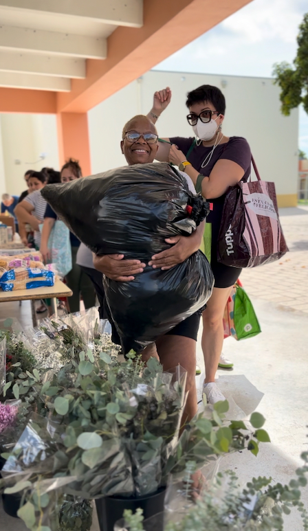 Woman smiles, holding large black trash bag with another woman raising an arm in celebration. Both are at a table with flowers, outside.