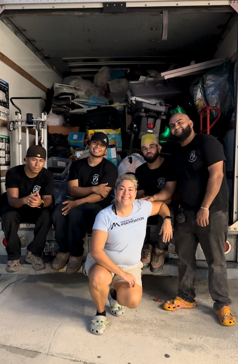 A group of six people smiling in front of a packed moving truck.  One person is kneeling, and the others are sitting or standing.