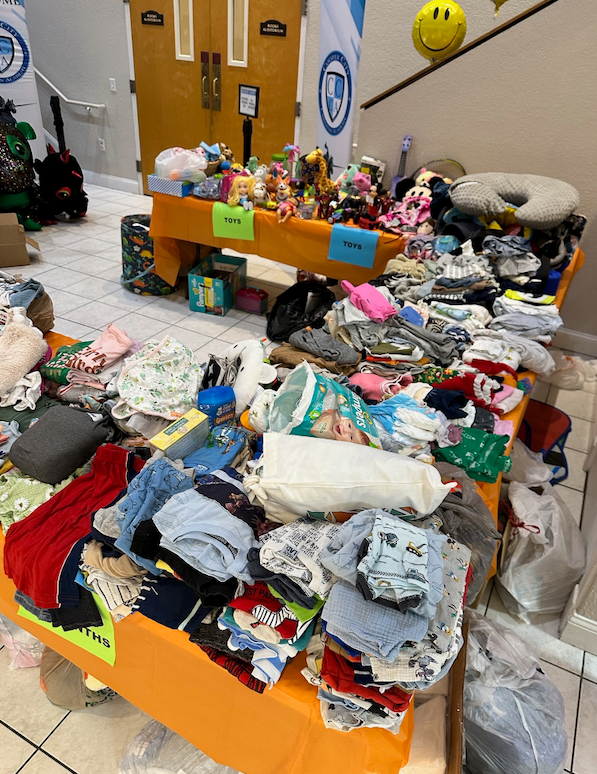 A table laden with baby clothes and toys in a brightly lit room, possibly a donation drive.