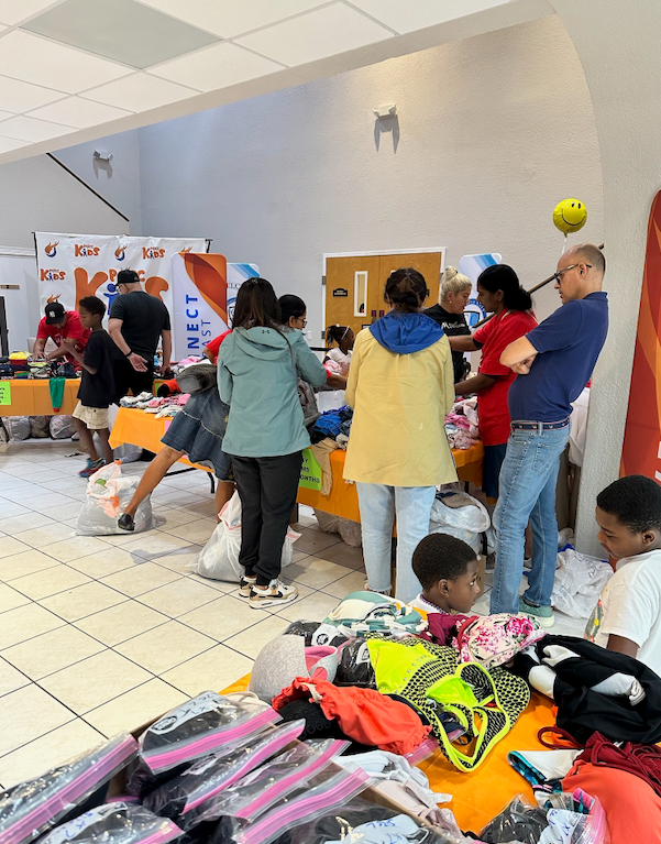 People browse and select items at a clothing giveaway in a brightly lit community center. Clothes are piled on tables, and volunteers assist shoppers.