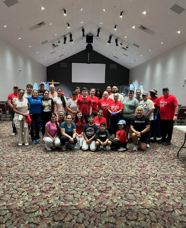 Group of people posing together in a church hall. Most wear red shirts. The room has beige carpet and a white screen.