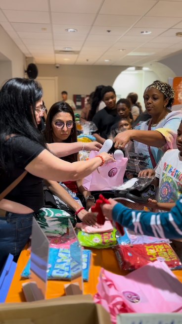 People at a table handing out pink bags filled with items. Inside a room, a line of people reaches the table.