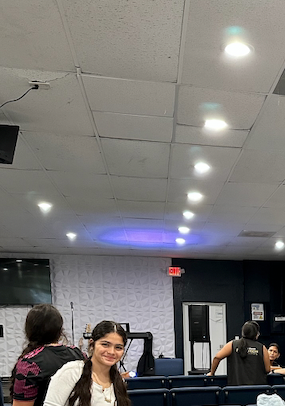 A young woman smiles in a brightly lit room with other people. White ceiling with recessed lights.