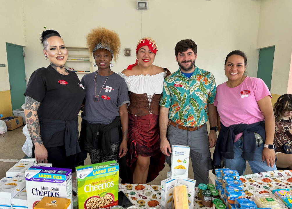 Five people smiling behind a table of food boxes, likely at a food drive or charity event. Setting is a brightly lit indoor space.