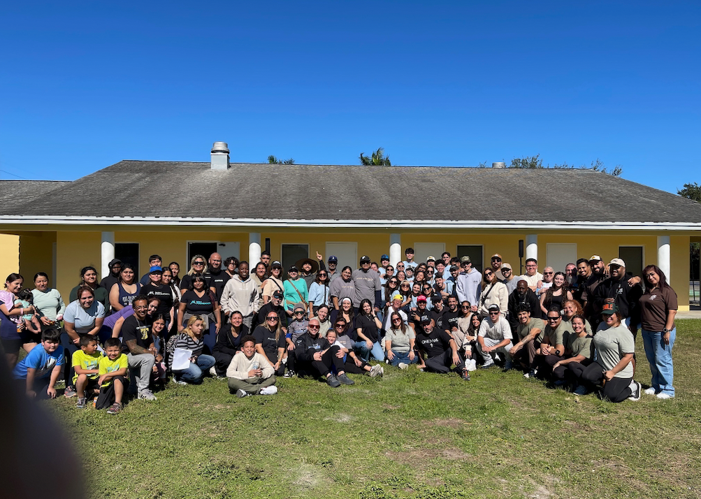 A large group of people pose for a photo in front of a yellow building on a sunny day. Most are wearing dark clothing.