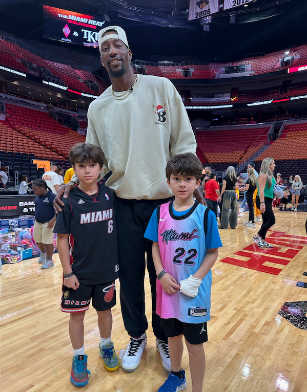 Basketball player Bam Adebayo with two young boys on a basketball court. Adebayo wears a sweatshirt; the boys wear jerseys.