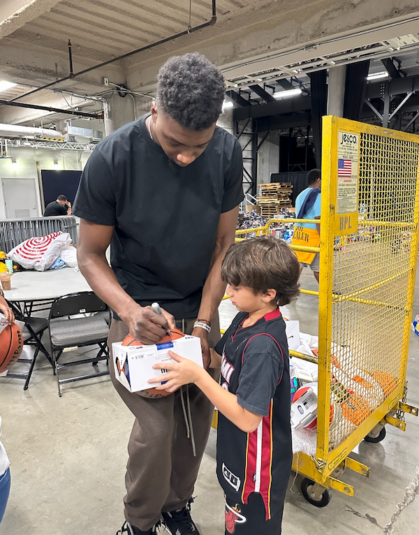 A basketball player signing a box for a young boy in a warehouse setting. The player is tall, wearing a black shirt and brown pants. The boy wears a basketball jersey.