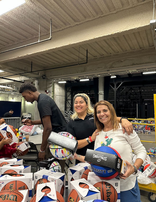 Three people pose with basketballs and boxes. A young man and two women smile, surrounded by sports equipment in a warehouse setting.