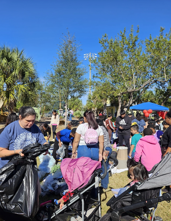 People gathered outdoors at a park, many sorting through items, likely clothing or donations. Several children in strollers are present on a sunny day.