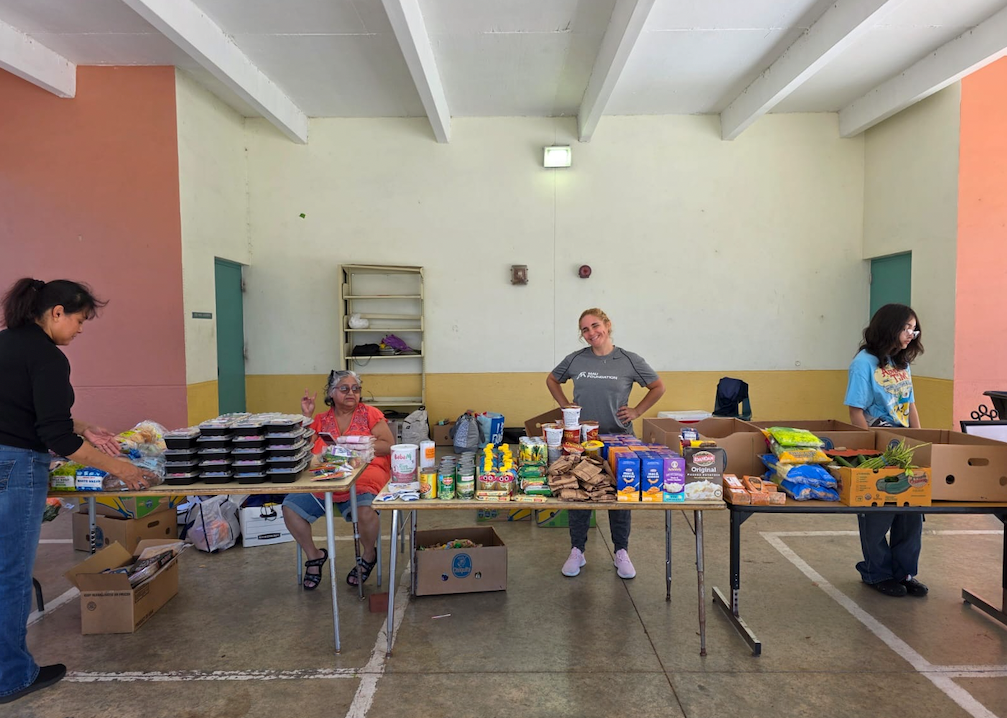 Volunteers at tables inside a building sort and package food items, including pre-made meals and snacks. The space is brightly lit.