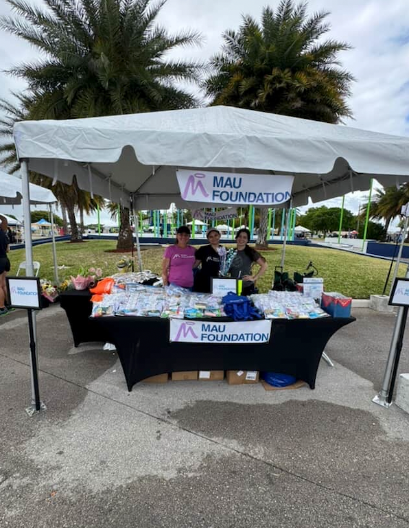 Three people stand behind a table with merchandise at a MAU Foundation booth under a white tent. The table is draped in black, set outdoors.