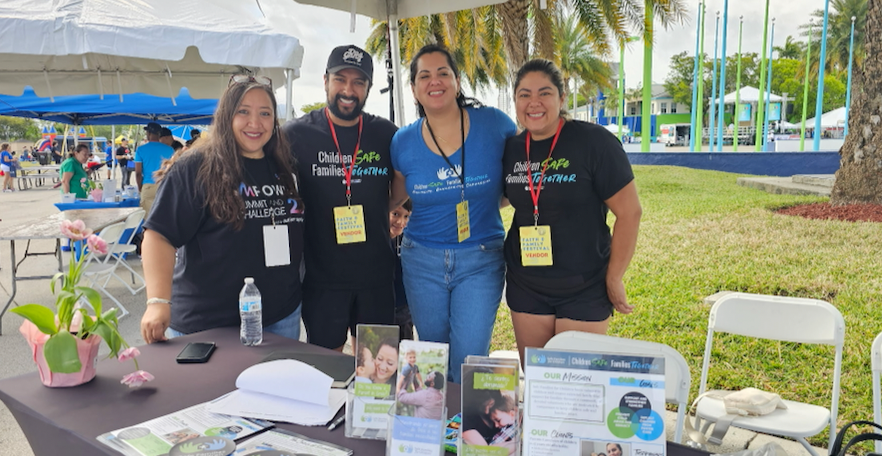 Four smiling people stand behind a table at an outdoor event. They wear matching shirts with a logo and name tags.
