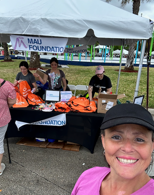 Woman smiling in selfie, in front of a MAU Foundation booth with volunteers. The booth has orange bags and merchandise.