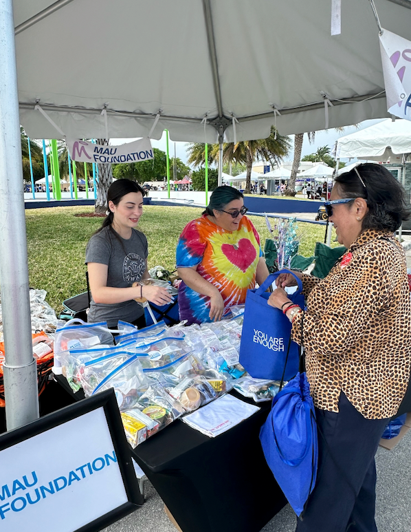 People at an outdoor booth: a vendor in tie-dye, a customer, and another vendor, smiling. The booth has products and a sign for the MAU Foundation.