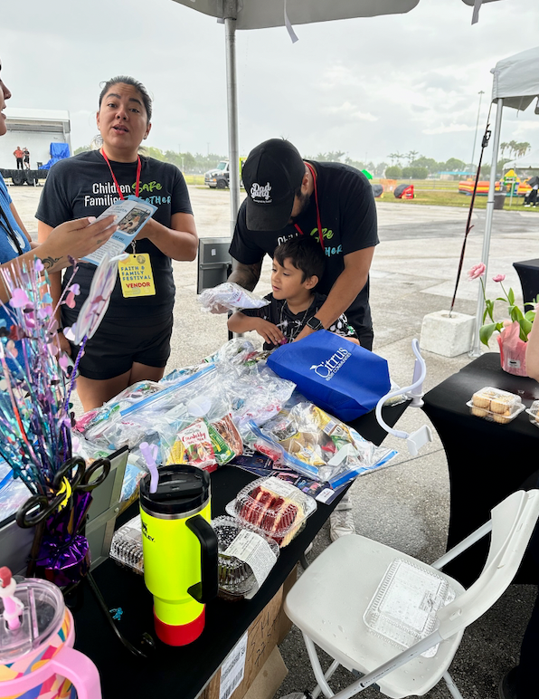 People at a table with food and drinks under a tent. A man and woman are attending to a child.