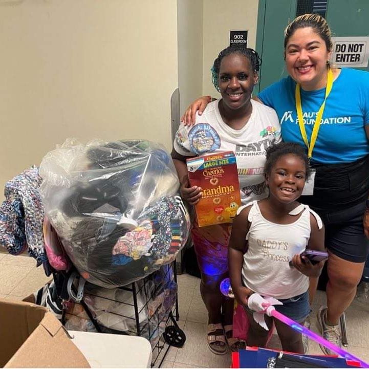 Four people, including a young girl, pose with donated items like clothing and a box of cereal inside a building.