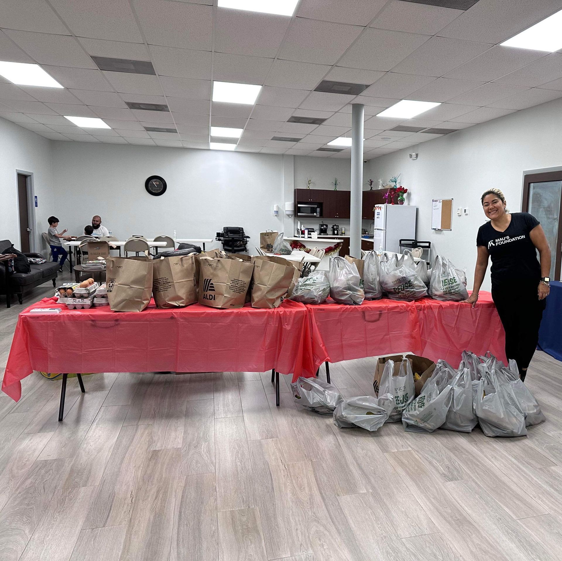 A woman stands beside a long table filled with bags of food, likely for distribution. Other people are in the background of a brightly lit room.