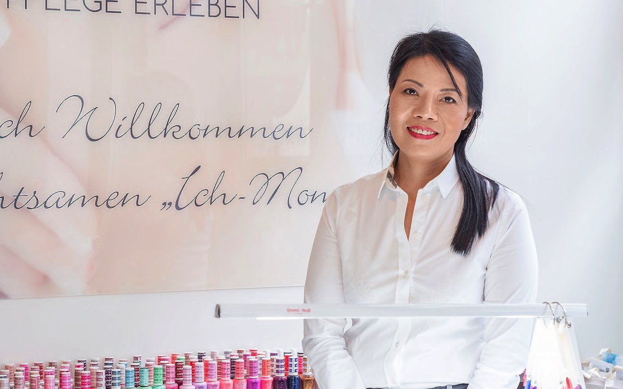 Die Betreiberin des Kosmetikstudios. Sie trägt ein weißes Hemd, hat dunkle Haare und steht vor dem Maniküretisch in ihrem Studio. 