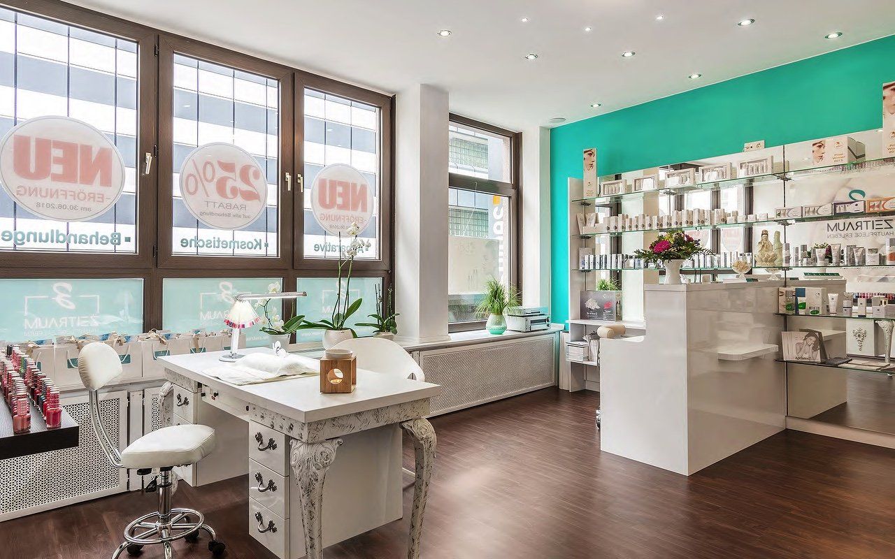 Innenraum eines Schönheitssalons mit Maniküretisch, weißem Empfangstresen und Regalen voller Produkte. Türkisfarbene Akzentwand.