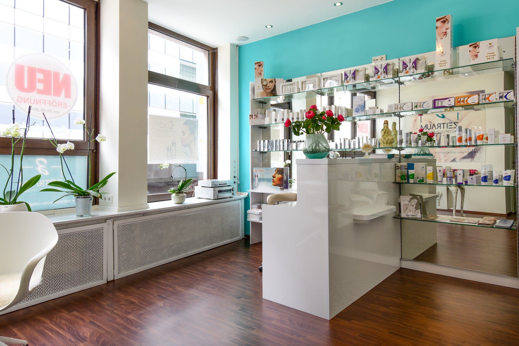 Empfangsbereich des Kosmetikstudios mit weißem Schreibtisch, blauer Wand, Produktregalen und großem Fenster.