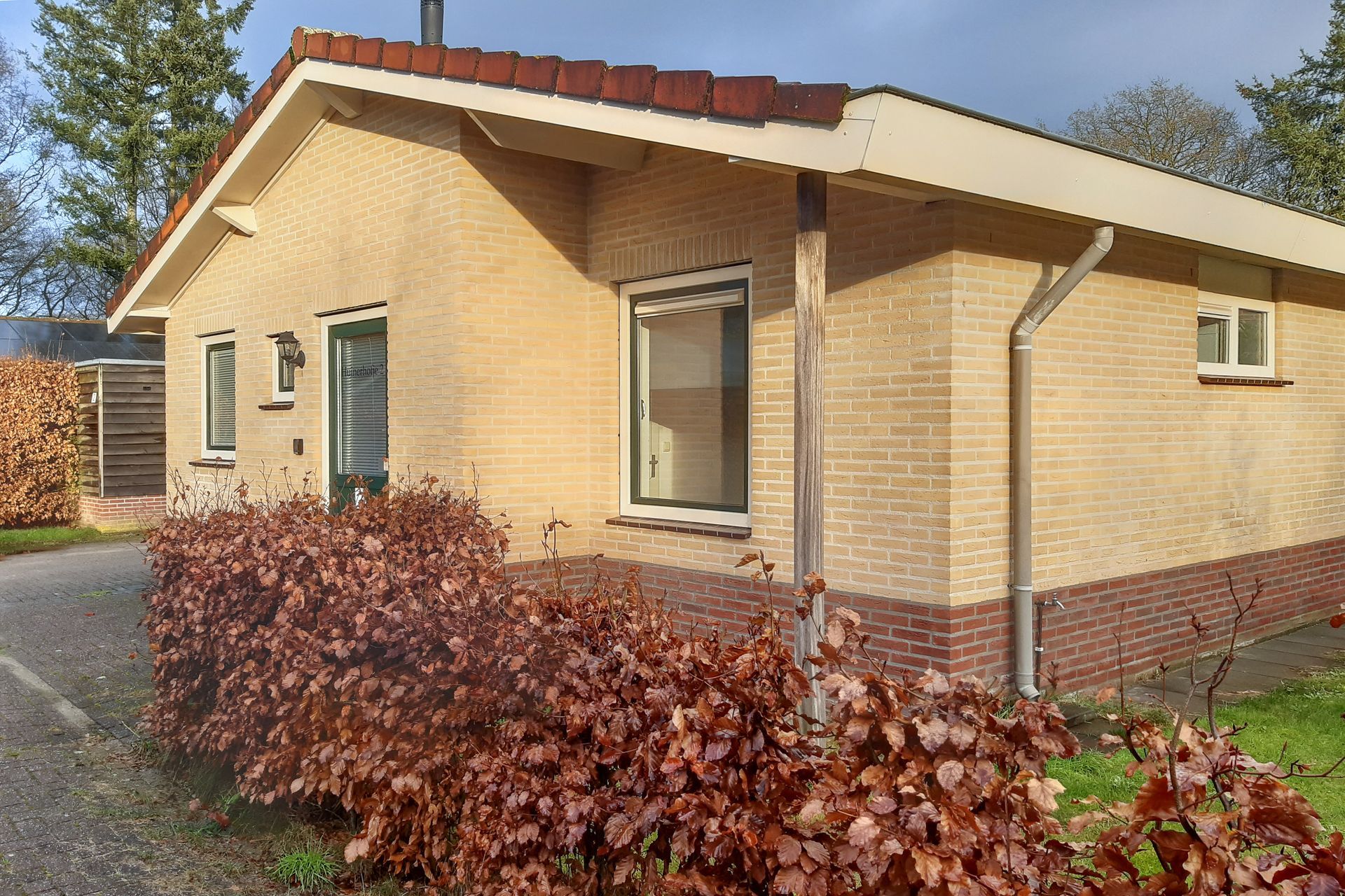 Een klein bakstenen huis met een rood pannendak, omgeven door struiken en bomen.