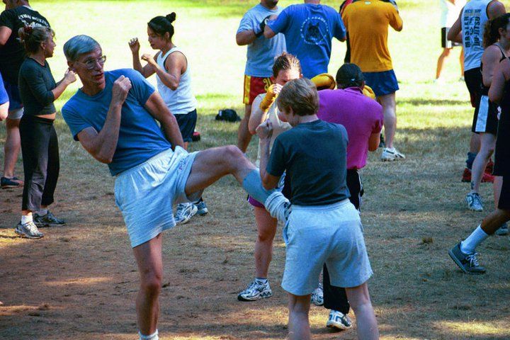 People practicing kickboxing in a park. Man in blue shirt kicks towards person holding pads; others in background.
