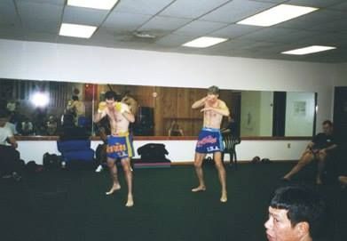Two men in Muay Thai stance, wearing blue shorts, in a gym with a mirror.