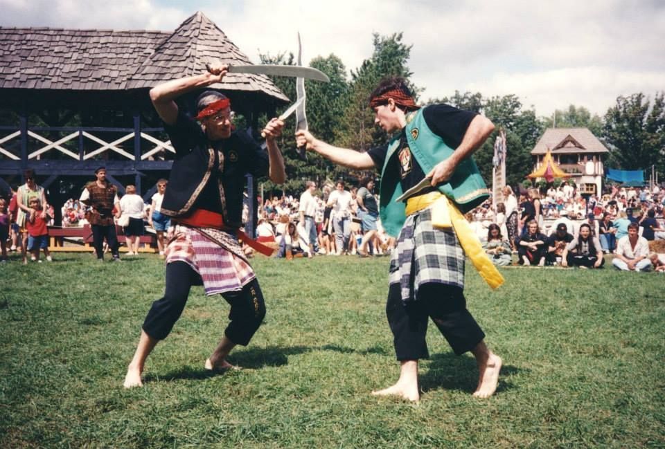 Two men practice sword fighting in an outdoor demonstration, wearing traditional clothing. People watch nearby.