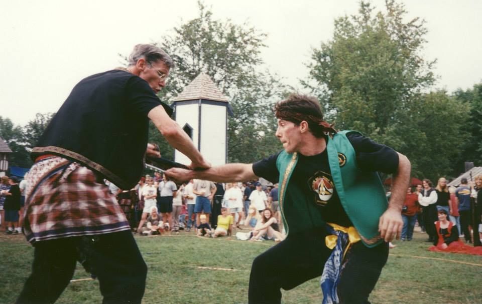 Two men in martial arts attire practice a move outdoors.