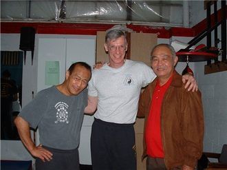 Three men posing together in a gym. Two Asian men flank a Caucasian man wearing a Yin Yang shirt.