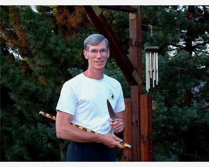Man holding a knife and staff outdoors, standing by a wooden structure with wind chimes, wearing a white shirt and dark pants.