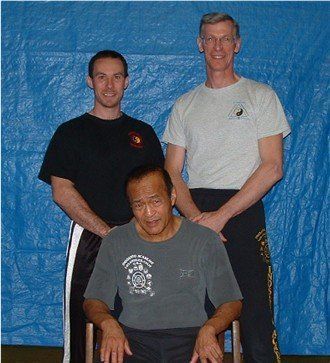 Three people in front of a blue backdrop: one seated in a chair, the other two standing. All are wearing t-shirts and dark pants.