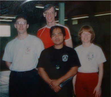Four people standing, two rows, in a training space. They wear t-shirts and pants.