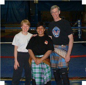 Three people standing in a boxing ring, wearing kilts and t-shirts, smiling.