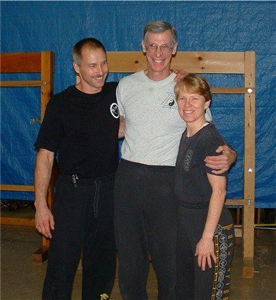 Three people, embracing. Center person taller than the others. All are wearing activewear in front of a blue backdrop.