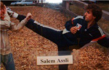 Man in blue tracksuit performing a high kick towards a person. Photo taken outdoors with leaves.
