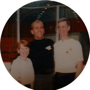 Three people posing together indoors; two men and a woman smiling near a window with a red wall.