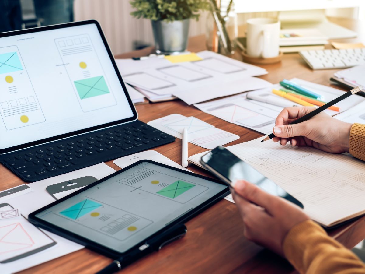 Une personne dessine des maquettes d'application à un bureau, avec un ordinateur portable, une tablette et un smartphone.