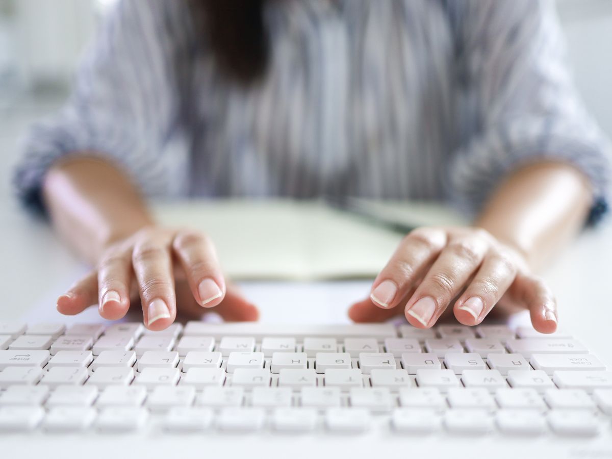 Mains tapant sur un clavier blanc, avec une personne floue et un cahier en arrière-plan.