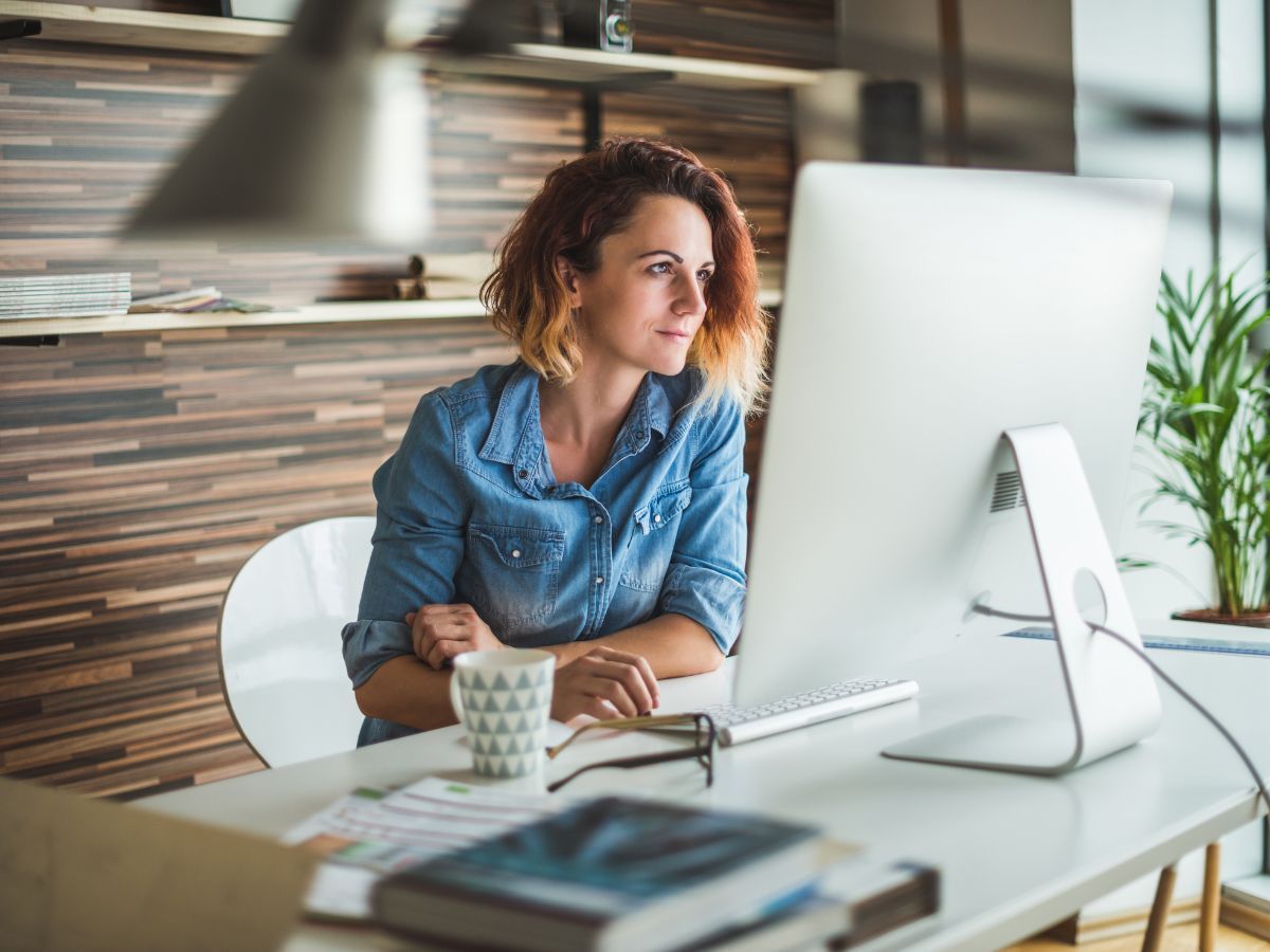 Femme en chemise en jean regardant un ordinateur, assise à son bureau, tasse de café à la main, bureau lumineux.